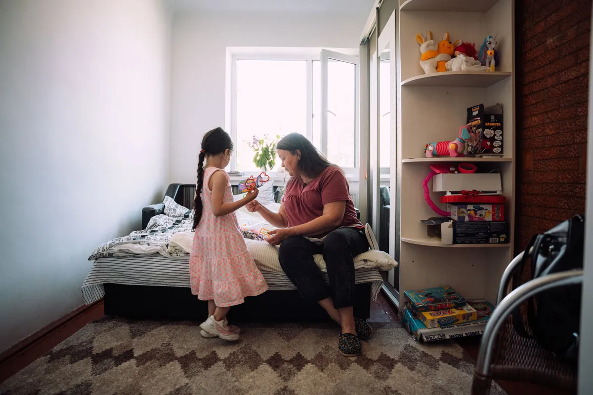 Mädchen mit Zöpfen im rosa Kleid zeigt einer Frau auf dem Bett ein Spielzeug, Regal mit Kuscheltieren, Flüchtlingsunterkunft in Moldau 2022, Reportage von Kilian Amrehn