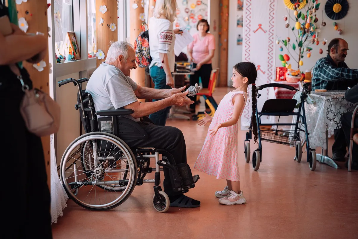 Älterer Mann im Rollstuhl reicht einem Mädchen im rosa Kleid ein Spielzeug, Gemeinschaftsraum, Flüchtlingsunterkunft in Moldau 2022, Reportage von Kilian Amrehn