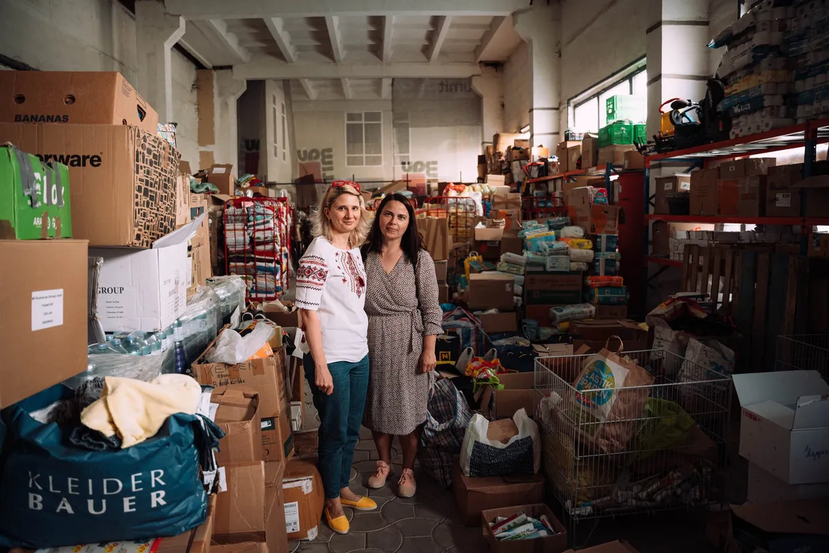 Zwei Frauen stehen in einer Lagerhalle zwischen gestapelten Spendenkartons und Taschen, Flüchtlingsunterkunft in Moldau 2022, Reportage von Kilian Amrehn