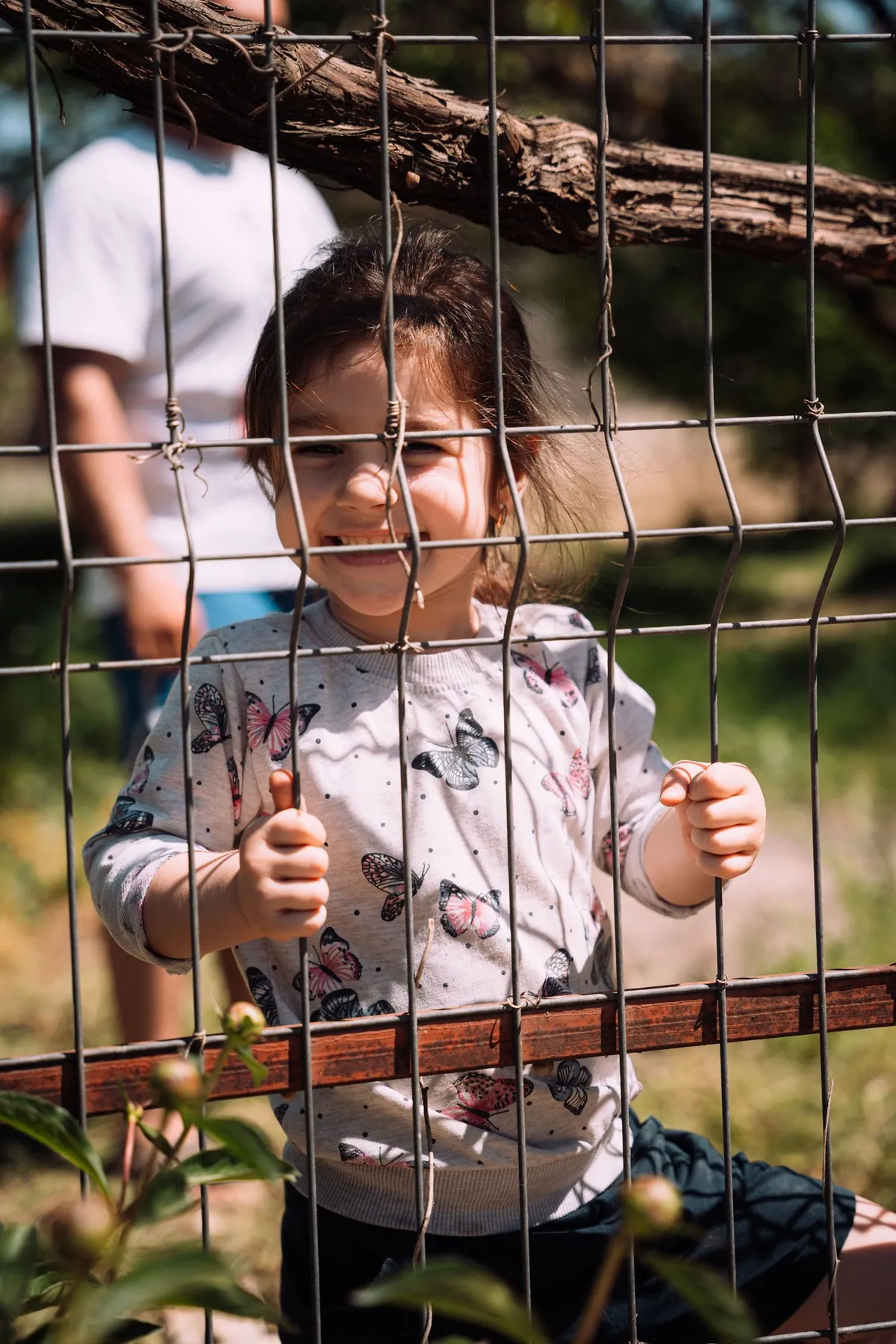 Kleines Mädchen im Schmetterlingspullover lächelt durch einen Maschendrahtzaun, Flüchtlingsunterkunft in Moldau 2022, Reportage von Kilian Amrehn