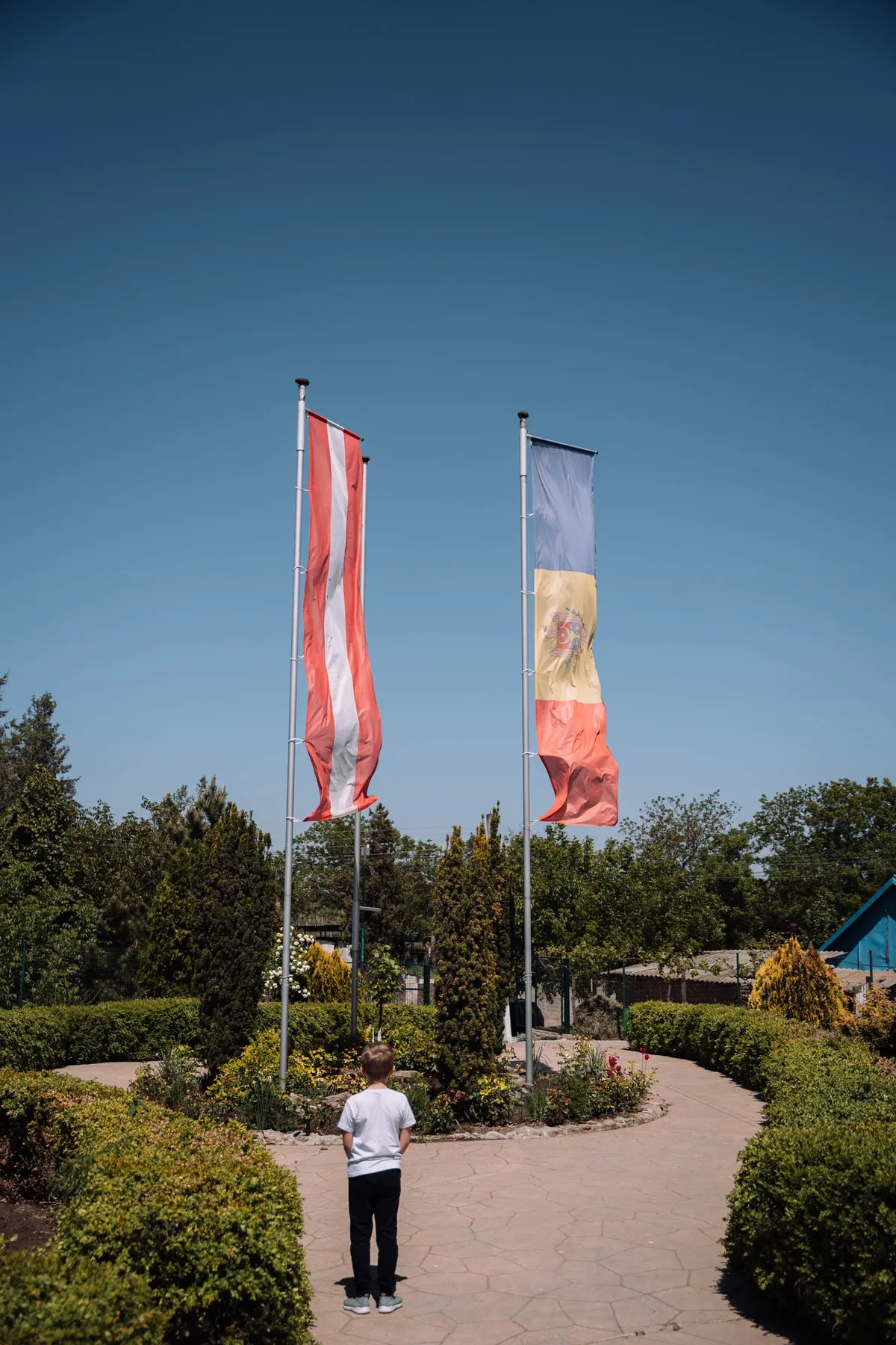 Junge steht in einem Garten mit Hecken vor zwei Flaggen an Masten, tiefblauer Himmel, Flüchtlingsunterkunft in Moldau 2022, Reportage von Kilian Amrehn
