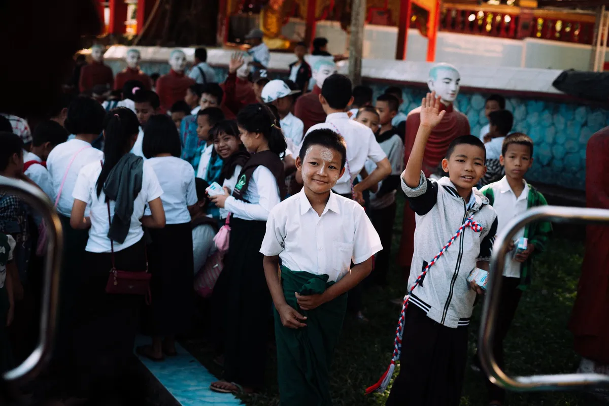 Schulkinder in weißen Hemden lächeln und winken in einer Gruppe vor einer Tempelanlage, Myanmar 2017, Reportage von Kilian Amrehn