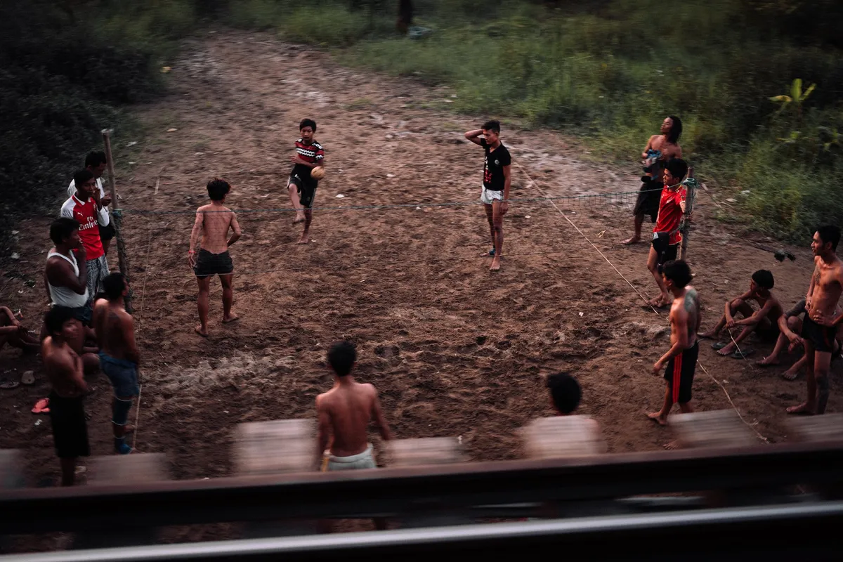 Jugendliche spielen auf einem lehmigen Platz mit einem Ball über ein Netz, Blick aus dem fahrenden Zug, Myanmar 2017, Reportage von Kilian Amrehn