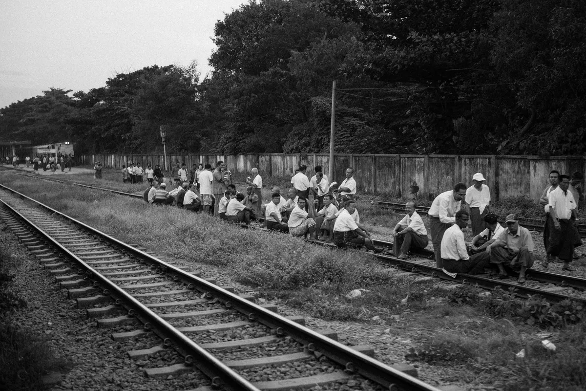 Gruppe von Männern in weißen Hemden sitzt neben Bahngleisen vor einer Mauer, schwarzweiß, Myanmar 2017, Reportage von Kilian Amrehn