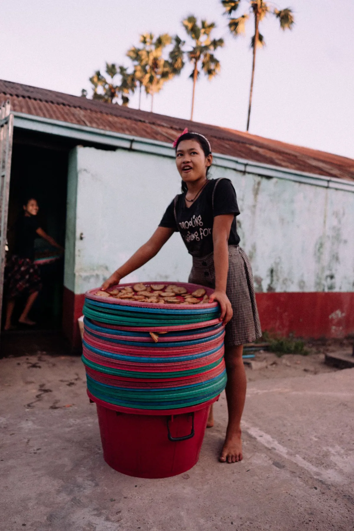 Junge Frau mit gestapelten bunten Plastikschüsseln vor einem Haus mit Wellblechdach, Palmen, Myanmar 2017, Reportage von Kilian Amrehn