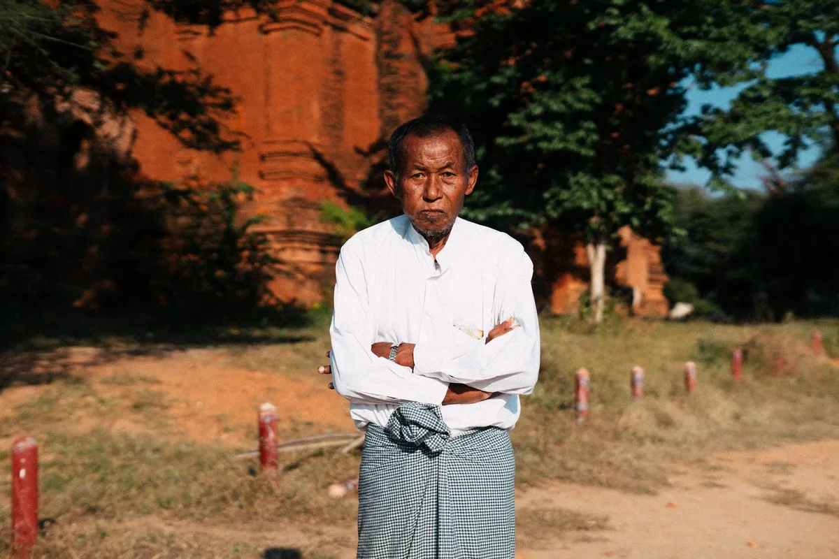 Älterer Mann in weißem Hemd und kariertem Wickelrock mit verschränkten Armen vor roter Backsteinruine, Myanmar 2017, Reportage von Kilian Amrehn
