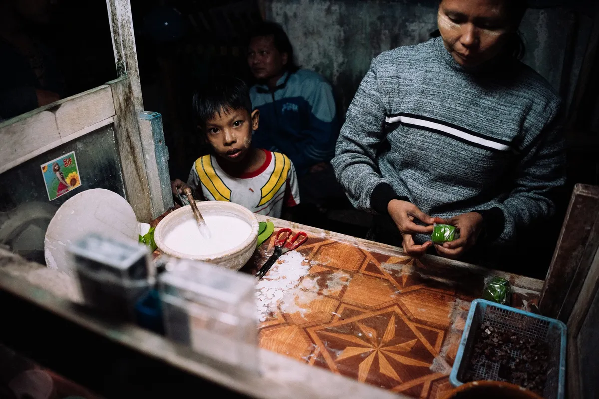 Frau bereitet an einem Kiosk kleine grüne Päckchen vor, Junge schaut in die Kamera, Lampenlicht, Myanmar 2017, Reportage von Kilian Amrehn