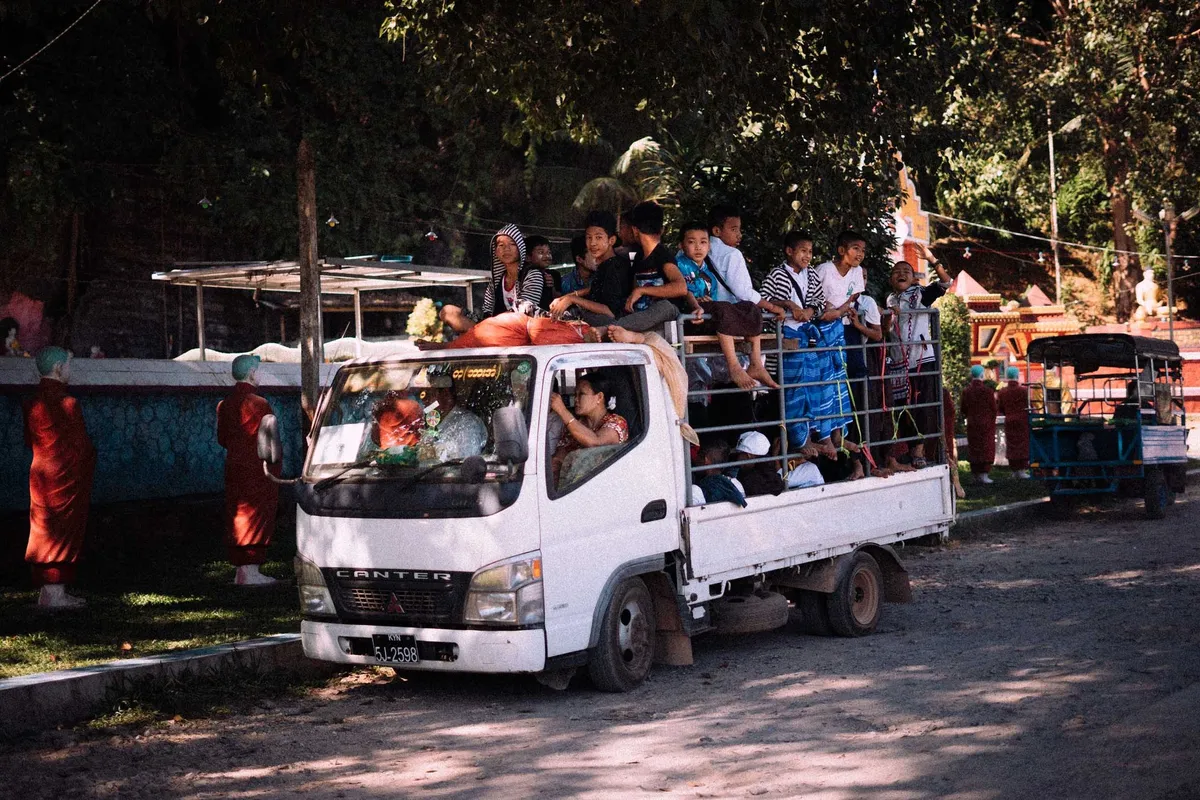 Menschen auf der Ladefläche eines weißen Kleinlasters im Schatten von Bäumen, Mönchsfiguren am Rand, Myanmar 2017, Reportage von Kilian Amrehn