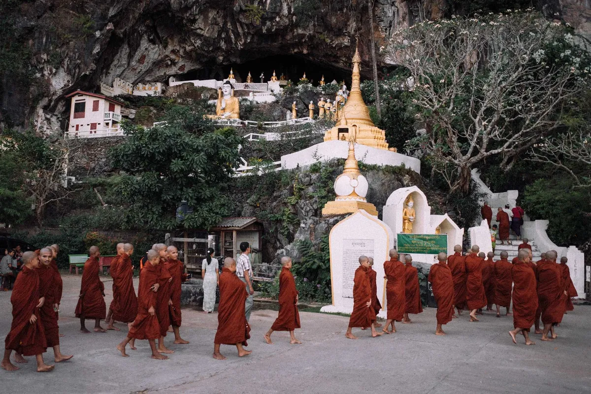 Mönche in roten Roben gehen an weißen Stupas vor einer Felswand mit goldener Pagode vorbei, Myanmar 2017, Reportage von Kilian Amrehn