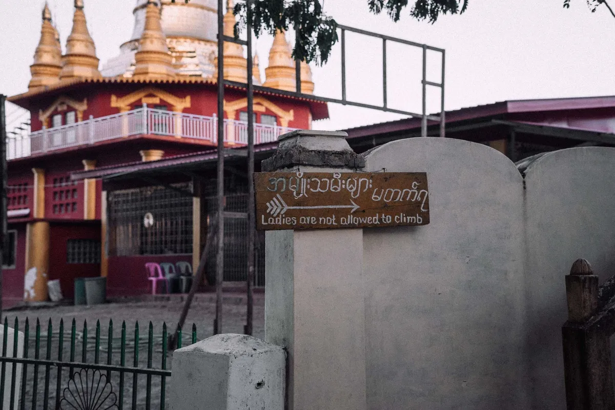 Schild Ladies are not allowed to climb an weißer Mauer vor rotem Tempel mit goldenen Spitzen, Myanmar 2017, Reportage von Kilian Amrehn