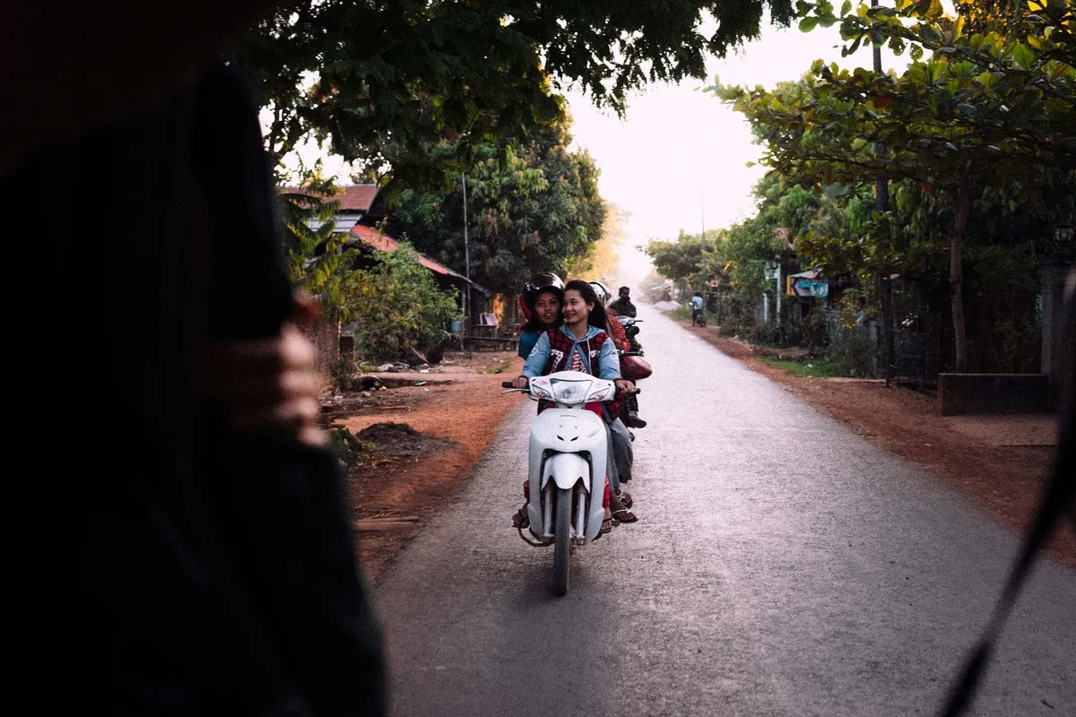 Lachende junge Frauen auf einem weißen Motorroller auf einer baumgesäumten Landstraße, Gegenlicht, Myanmar 2017, Reportage von Kilian Amrehn