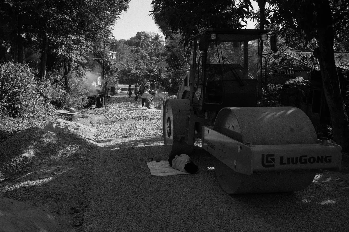 Arbeiter ruht im Schatten neben einer Straßenwalze auf einem Schotterweg zwischen Bäumen, schwarzweiß, Myanmar 2017, Reportage von Kilian Amrehn