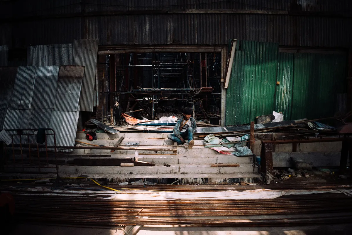 Junger Mann sitzt auf Treppenstufen in einer Halle aus Wellblech und Bauschutt, Lichtstreifen, Thailand 2017, Reportage von Kilian Amrehn