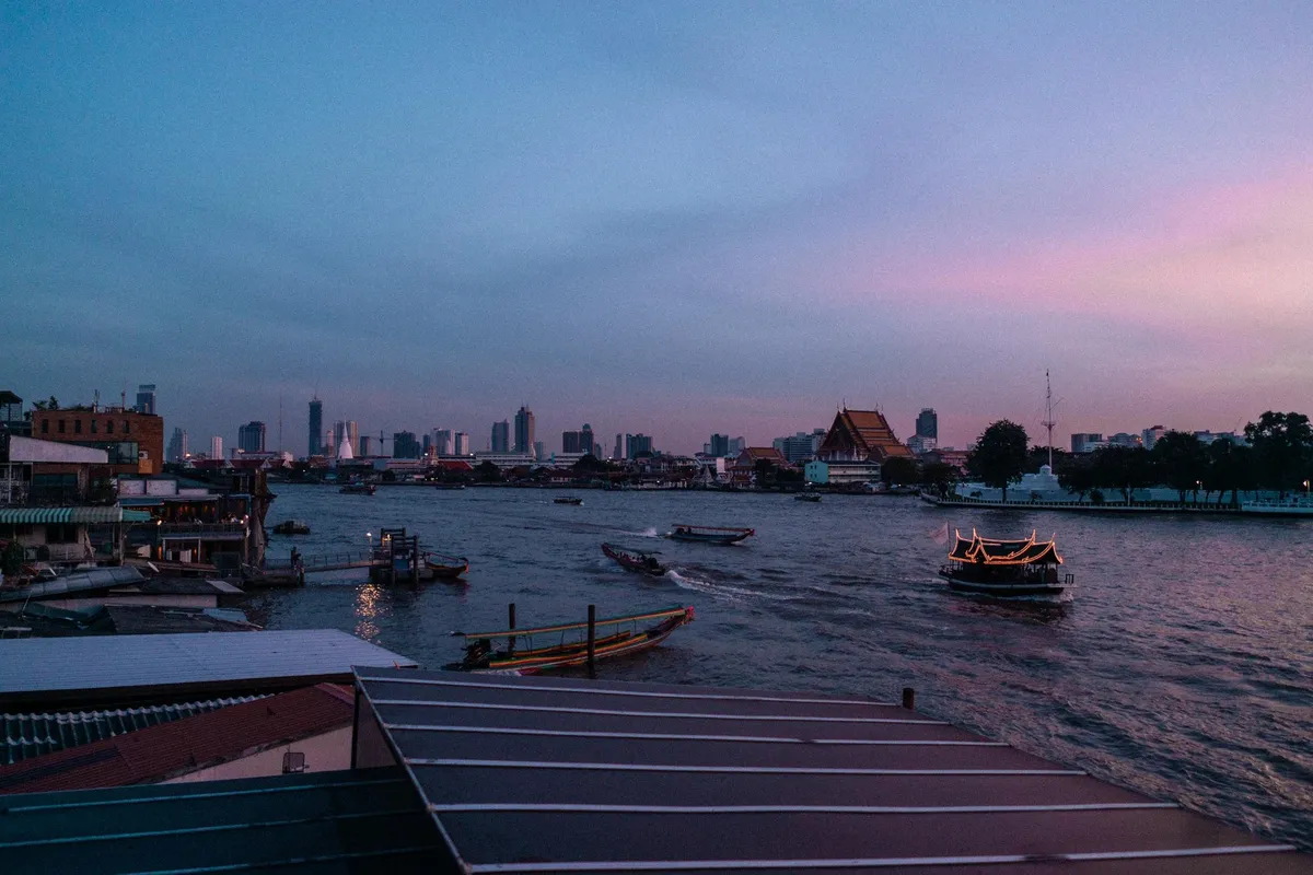 Fluss mit Booten in der Abenddämmerung vor Skyline und Tempeldach, rosa Himmel, Thailand 2017, Reportage von Kilian Amrehn
