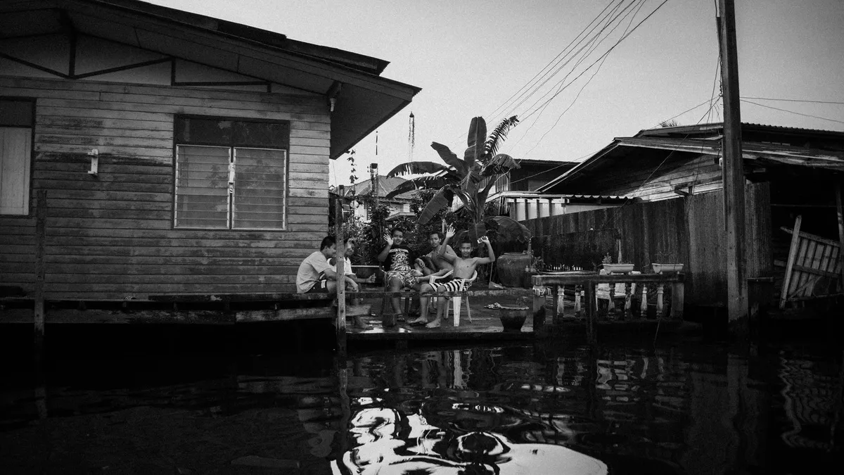 Familie sitzt auf der Holzterrasse eines Stelzenhauses am Kanal, Spiegelung im Wasser, schwarzweiß, Thailand 2017, Reportage von Kilian Amrehn