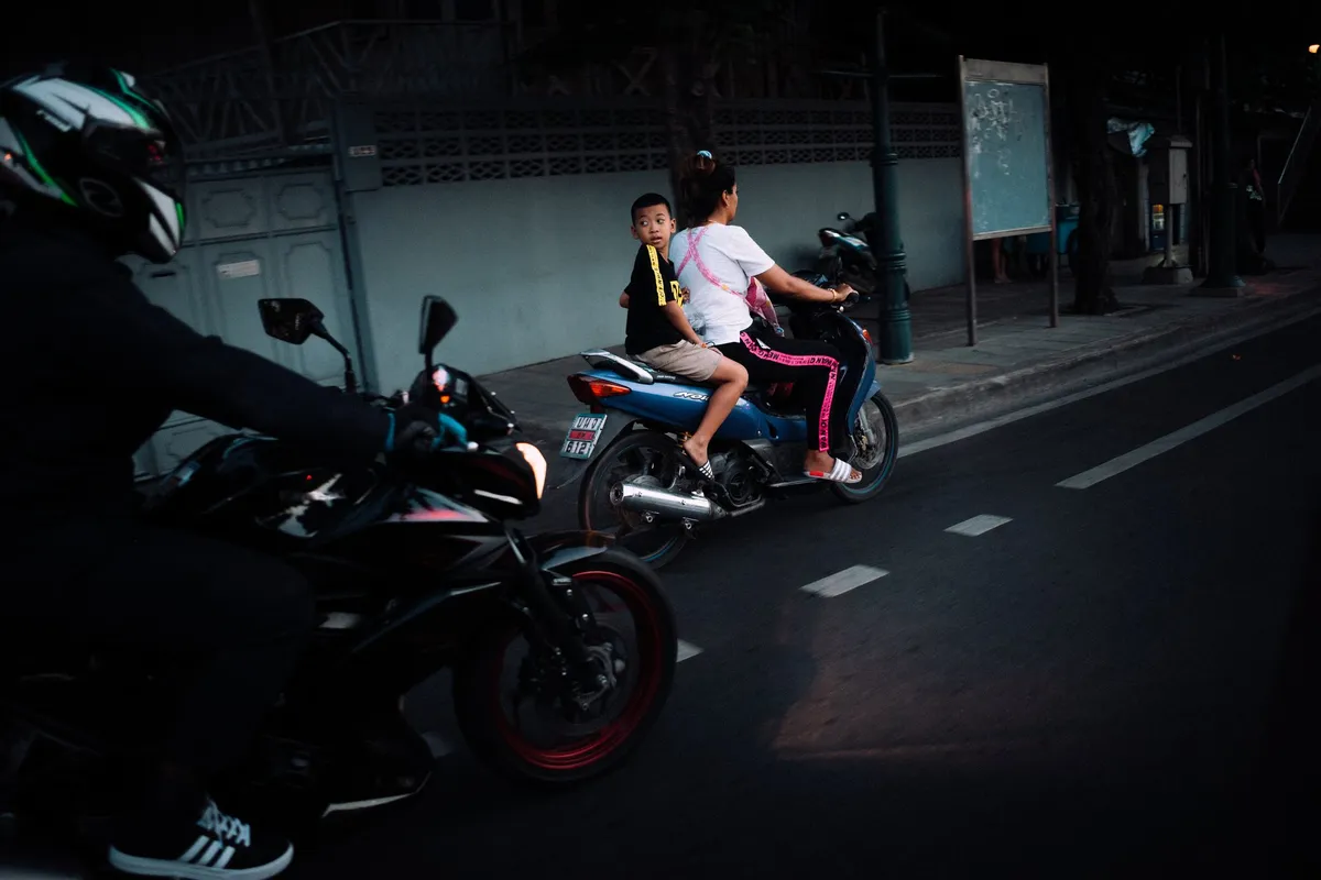 Frau und Kind auf einem Motorroller neben einem Motorradfahrer mit Helm, Straße in der Dämmerung, Thailand 2017, Reportage von Kilian Amrehn
