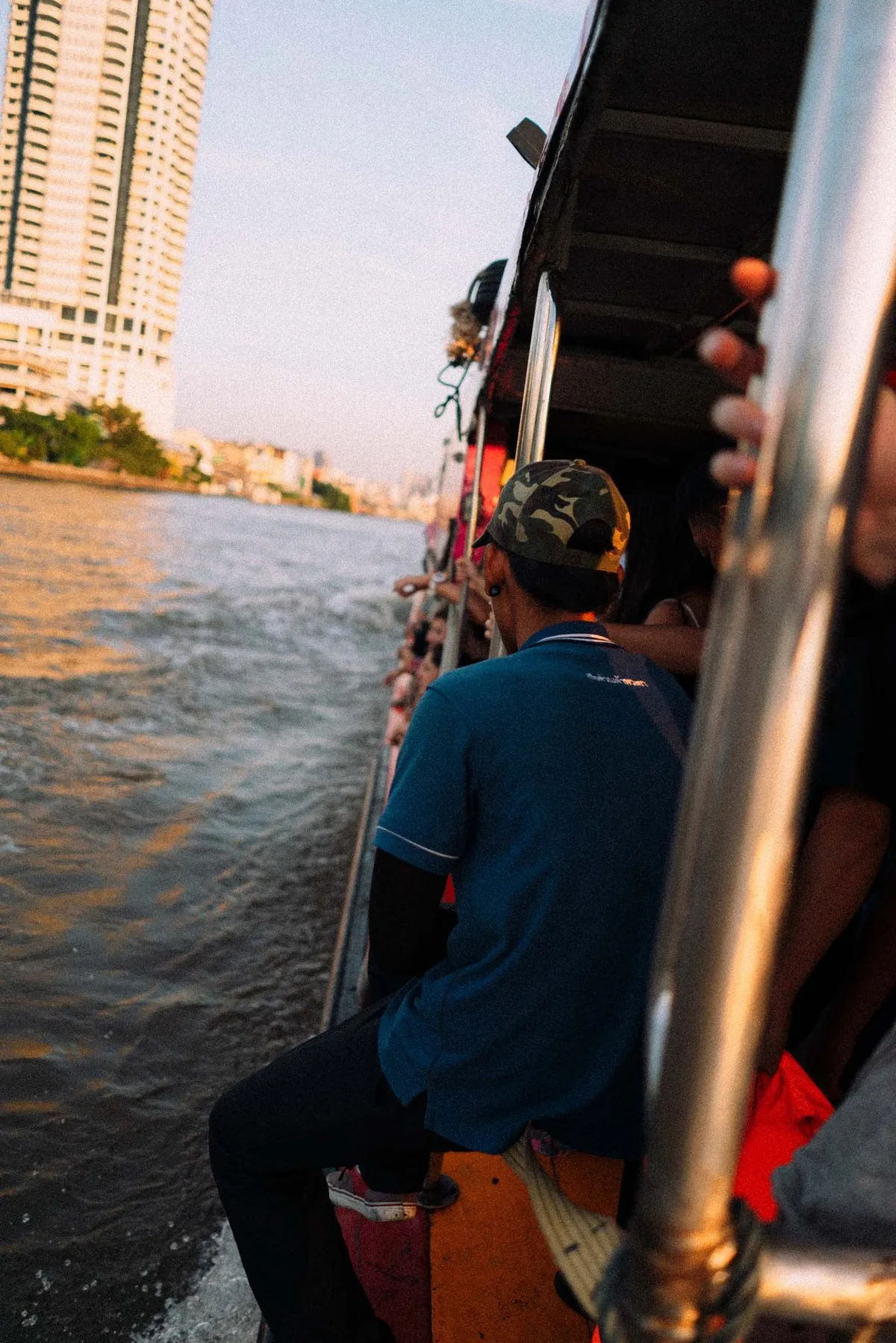 Mann mit Kappe sitzt am Rand eines fahrenden Flussboots, Hochhaus und Abendsonne, Thailand 2017, Reportage von Kilian Amrehn