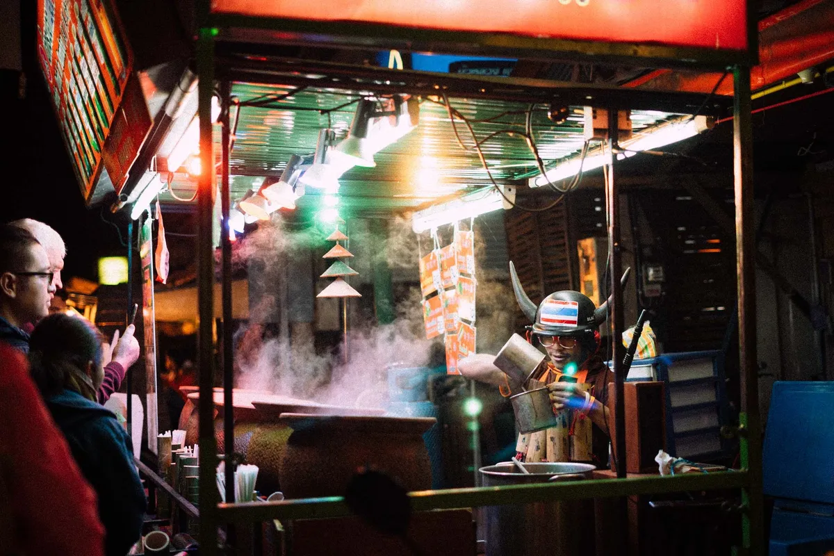 Garküche bei Nacht mit Dampf und bunten Lichtern, Koch mit Wikingerhelm in Flaggenfarben, Thailand 2017, Reportage von Kilian Amrehn