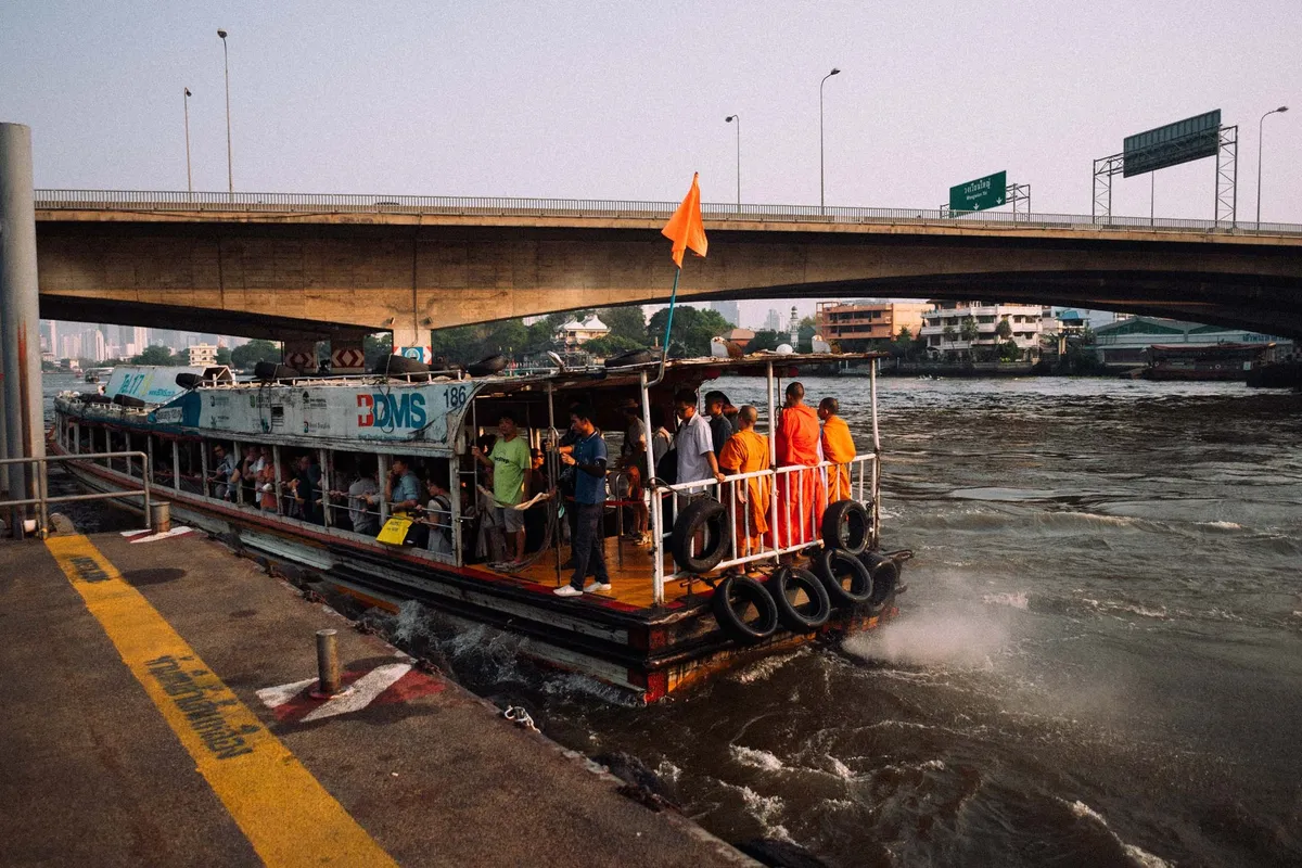 Voll besetzte Fähre mit Mönchen in orangen Roben legt unter einer Brücke am Anleger an, Thailand 2017, Reportage von Kilian Amrehn