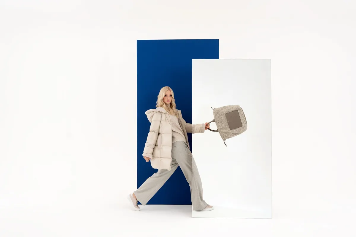 AppelrathCüpper, blonde model in a light puffer jacket swinging a knit handbag before a blue panel and mirror, campaign 2022, photographed by Kilian Amrehn