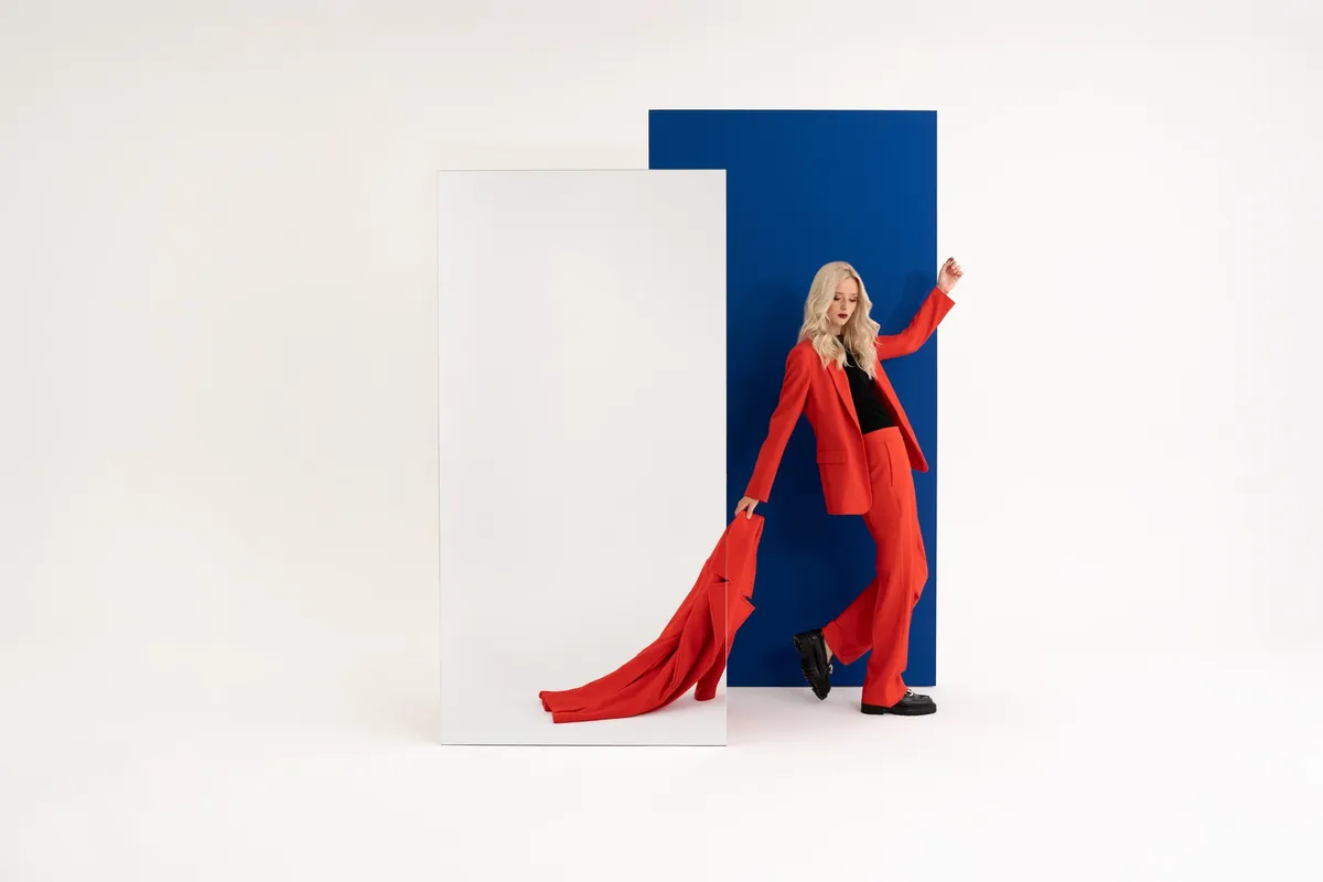 AppelrathCüpper, blonde model in a red trouser suit trailing a red coat, blue panel and mirror, campaign 2022, photographed by Kilian Amrehn
