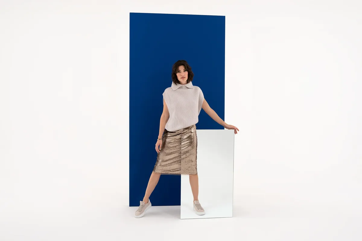 AppelrathCüpper, model with short hair in a pale knit slipover and gold sequin skirt before a blue panel and mirror, campaign 2022, photographed by Kilian Amrehn
