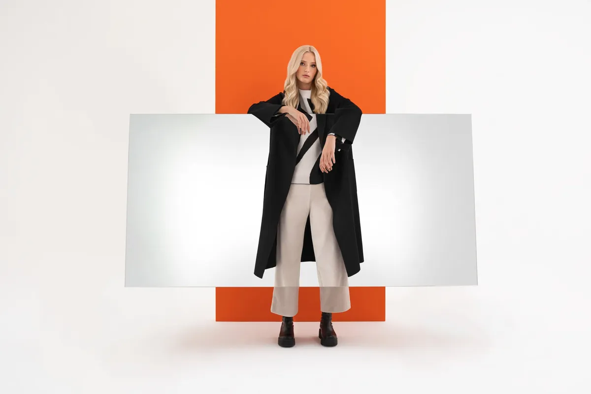 AppelrathCüpper, blonde model in a long black coat and light trousers behind a horizontal mirror before an orange panel, campaign 2022, photographed by Kilian Amrehn