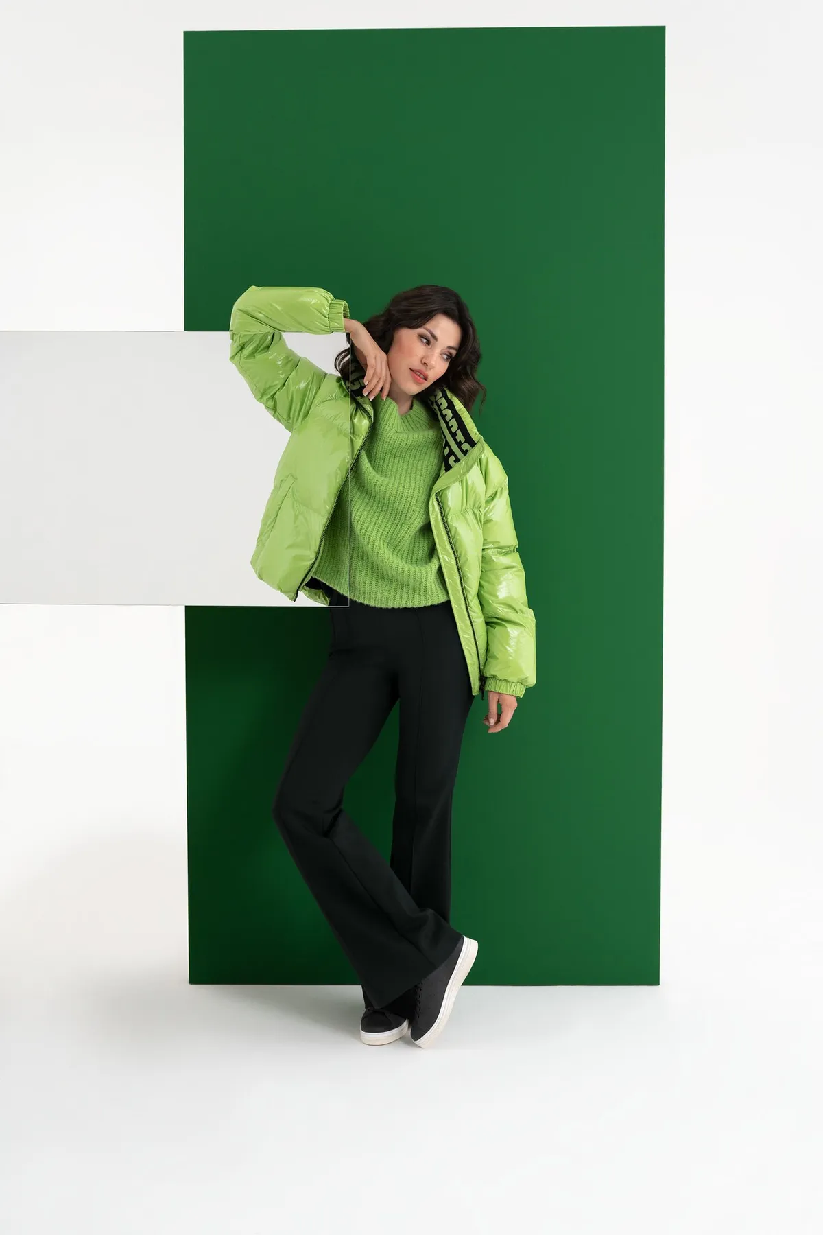 AppelrathCüpper, model in a lime glossy puffer jacket and knit sweater leaning against a dark green panel, campaign 2022, photographed by Kilian Amrehn