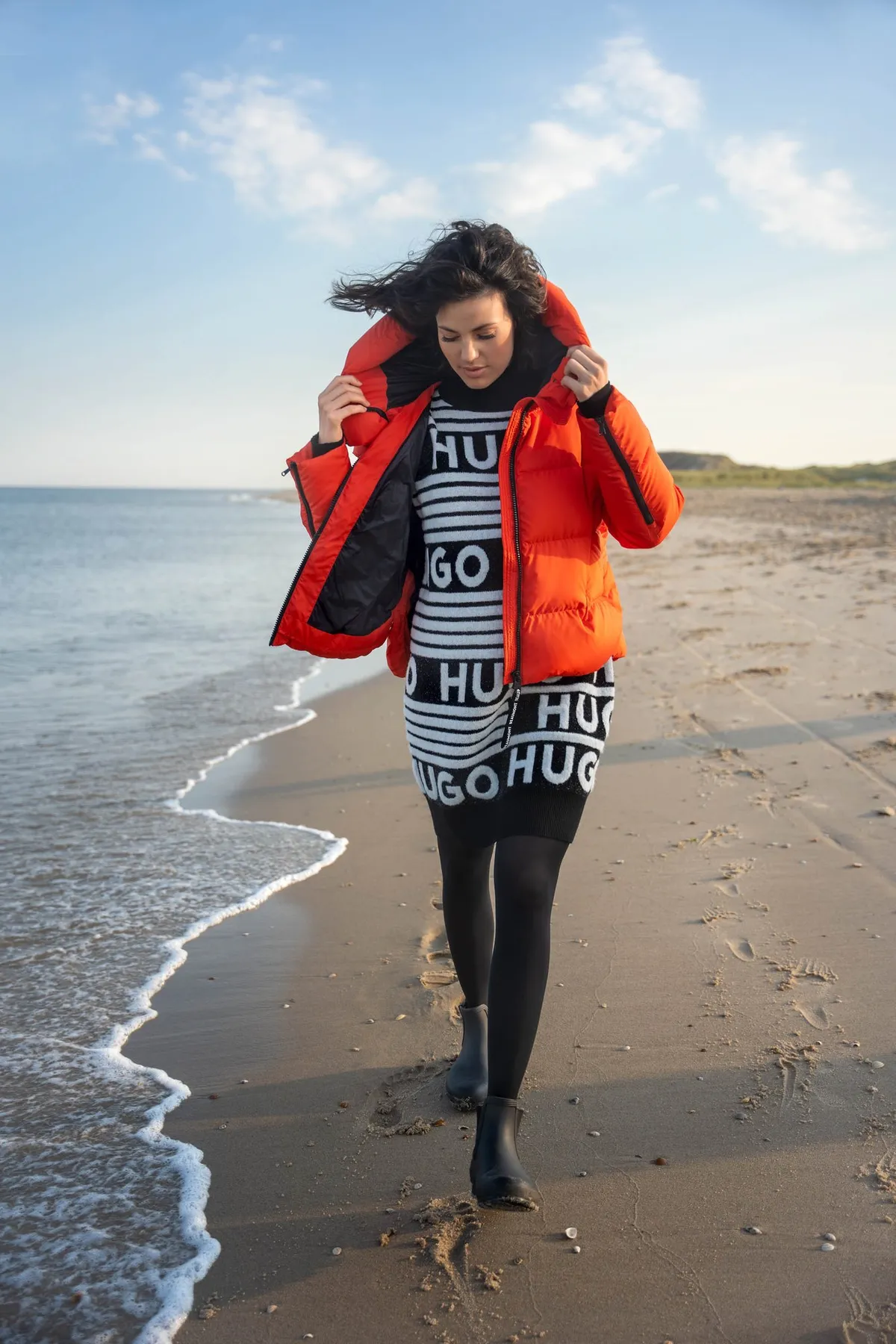 AppelrathCüpper, model in an orange puffer jacket and HUGO logo knit dress walking along the shoreline, campaign 2022, photographed by Kilian Amrehn