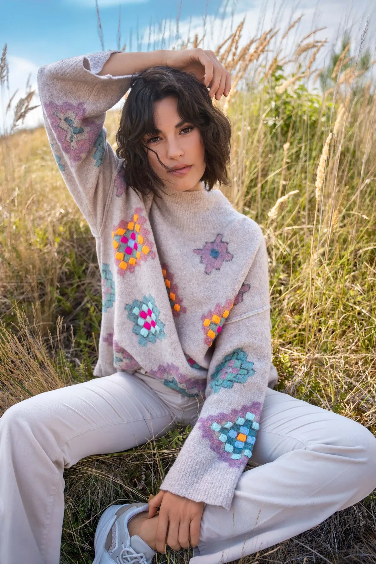 AppelrathCüpper, model in a sweater with colourful granny squares and white trousers sitting in tall dune grass, campaign 2022, photographed by Kilian Amrehn