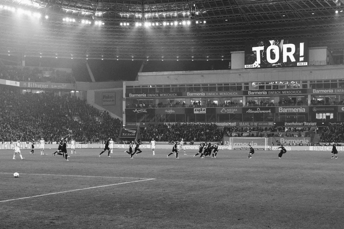 Bundesliga, view across the pitch under floodlights, scoreboard reading TOR, packed stands in black and white, sports photography, photographed by Kilian Amrehn