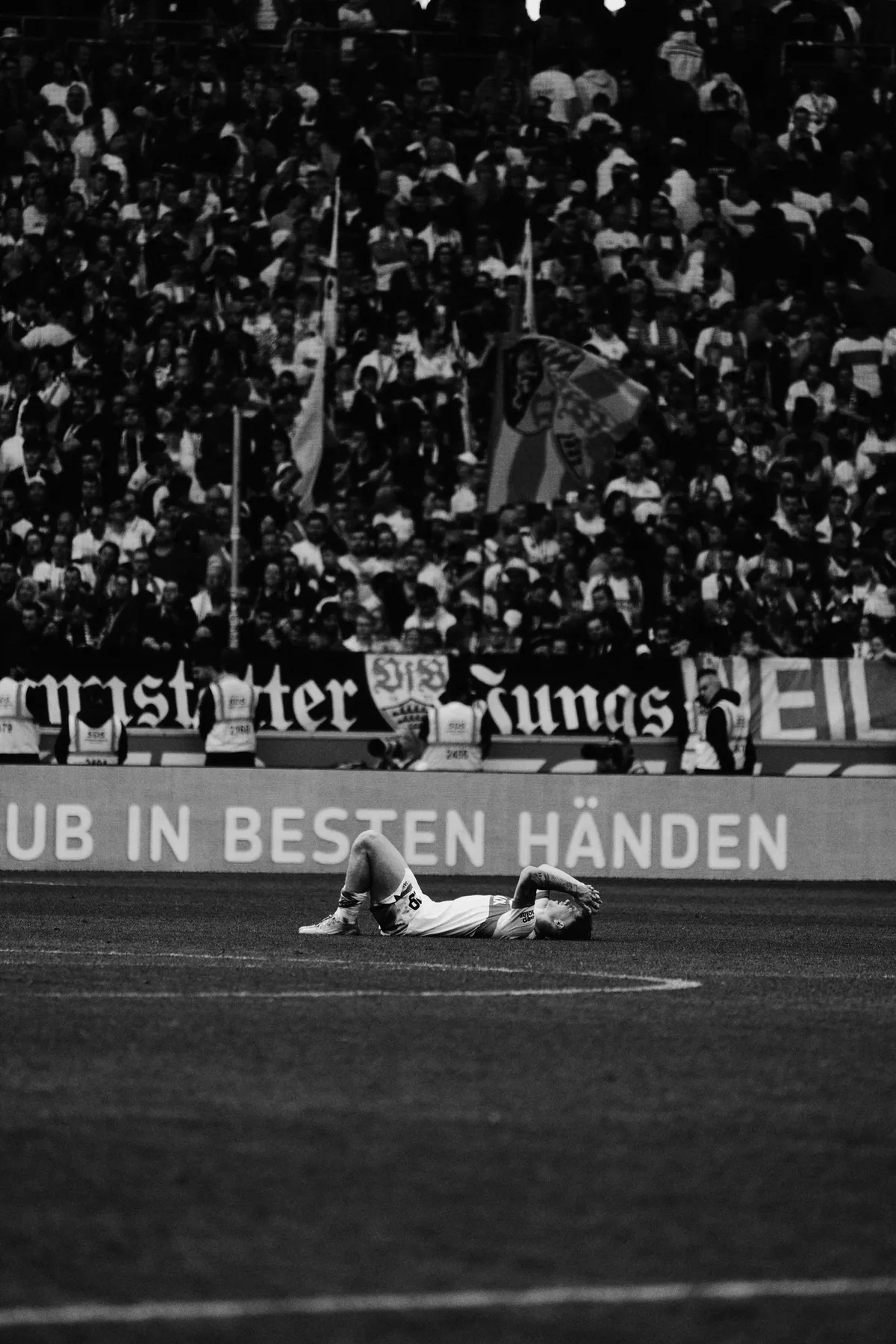Bundesliga, player lying alone on the pitch in front of the dark fan stand, black and white, sports photography, photographed by Kilian Amrehn
