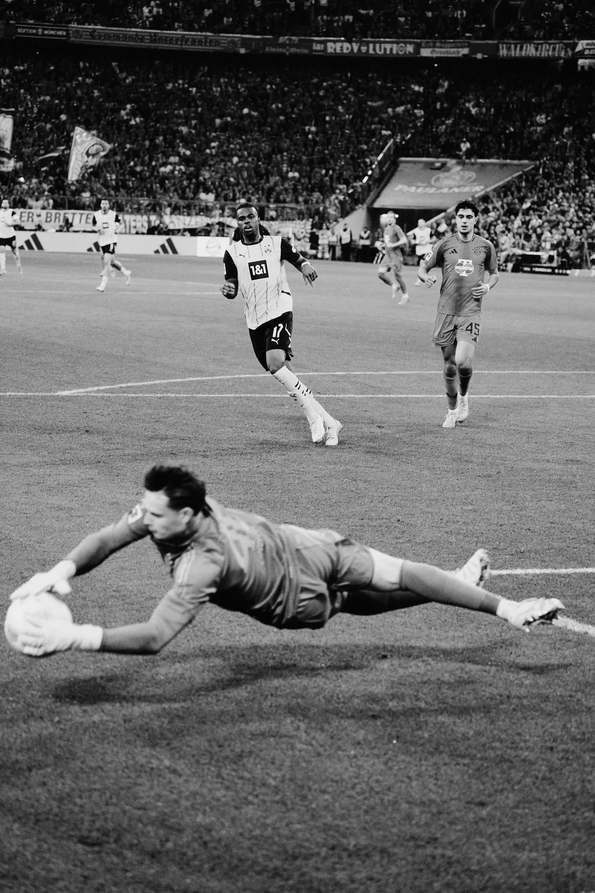 Bundesliga, goalkeeper diving low for the ball as a striker runs in behind, black and white, sports photography, photographed by Kilian Amrehn