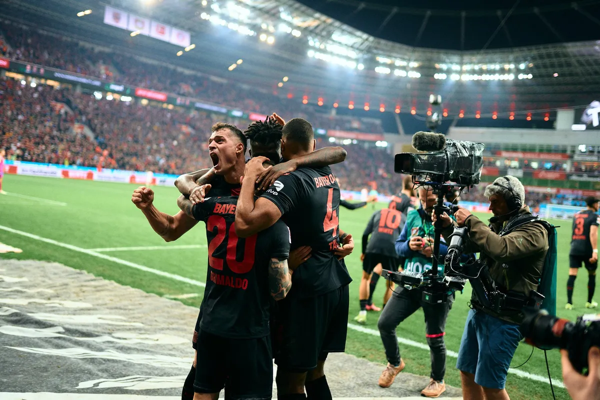 Bundesliga, Bayer 04 players celebrating in an embrace at the touchline as a TV cameraman films, floodlights and red stands, sports photography, photographed by Kilian Amrehn