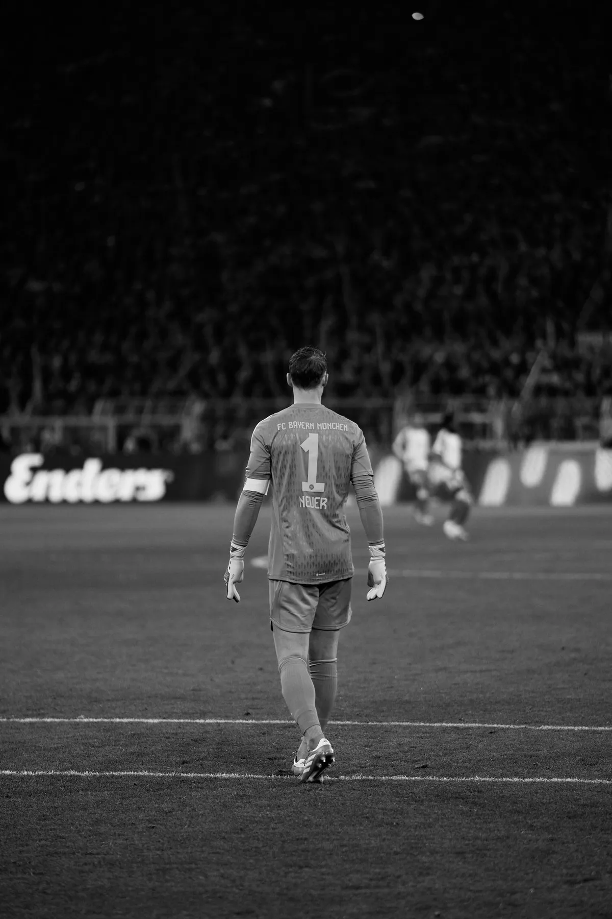 Bundesliga, goalkeeper wearing number 1 walking alone across the pitch before a dark stand, back view in black and white, sports photography, photographed by Kilian Amrehn