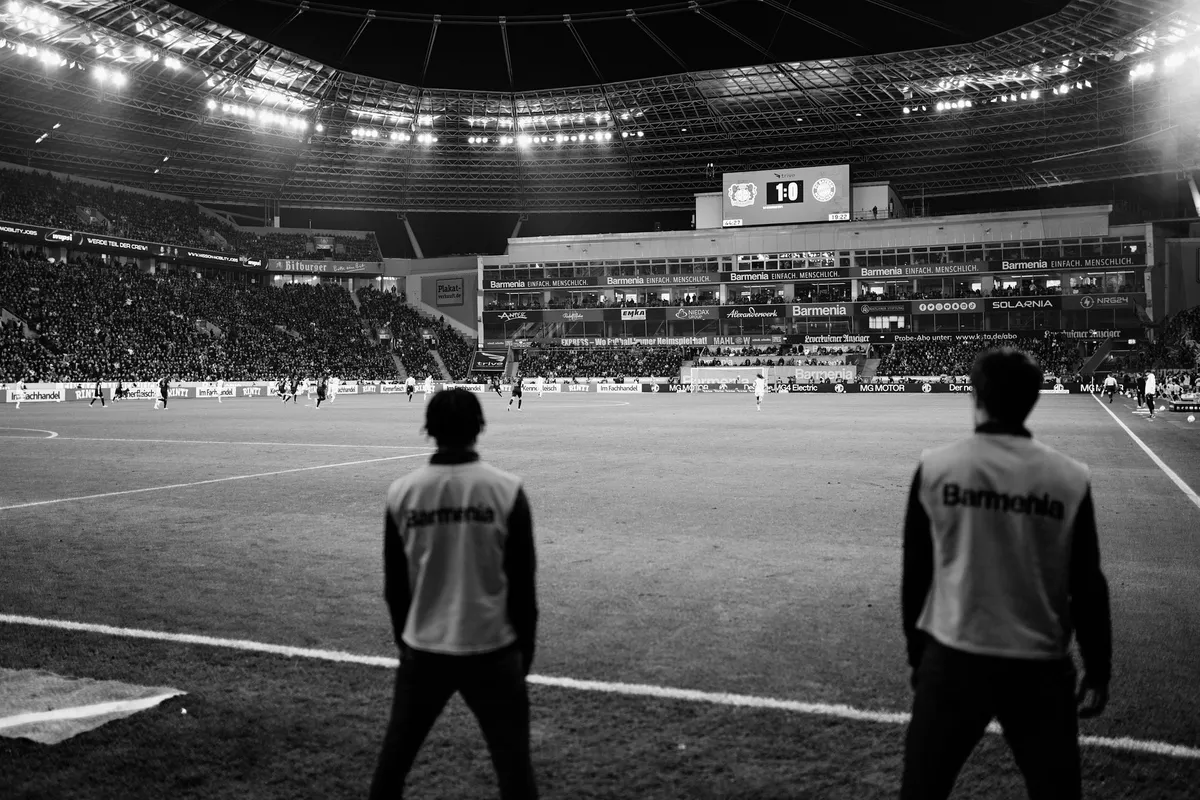 Bundesliga, two staff in bibs standing at the touchline of a floodlit stadium, scoreboard 1:0, black and white, sports photography, photographed by Kilian Amrehn