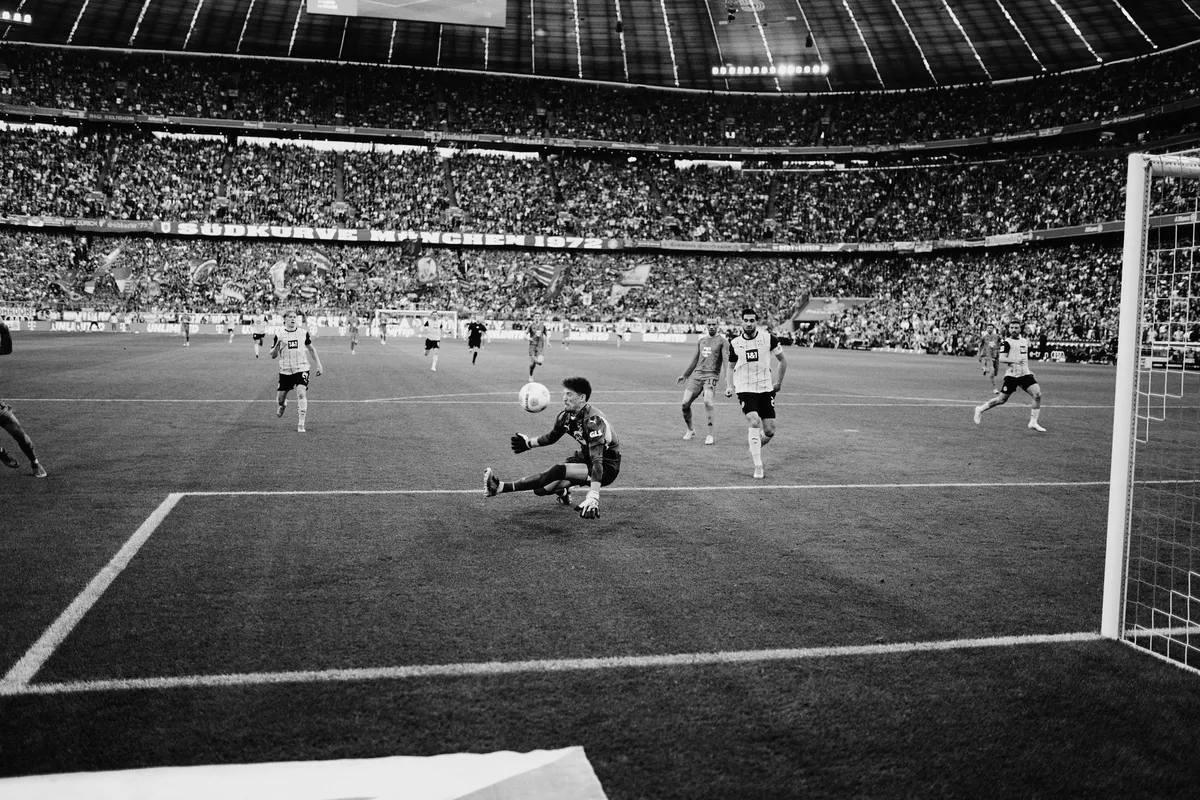 Bundesliga, goalkeeper diving for the ball in the box in front of packed stands, wide angle in black and white, sports photography, photographed by Kilian Amrehn