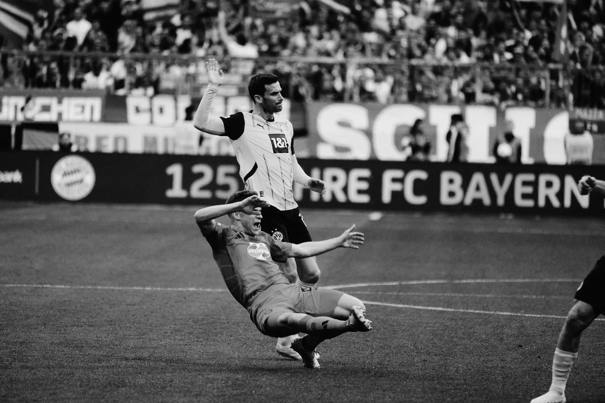 Bundesliga, challenge with a player falling in front of a 125 years FC Bayern board, black and white, sports photography, photographed by Kilian Amrehn