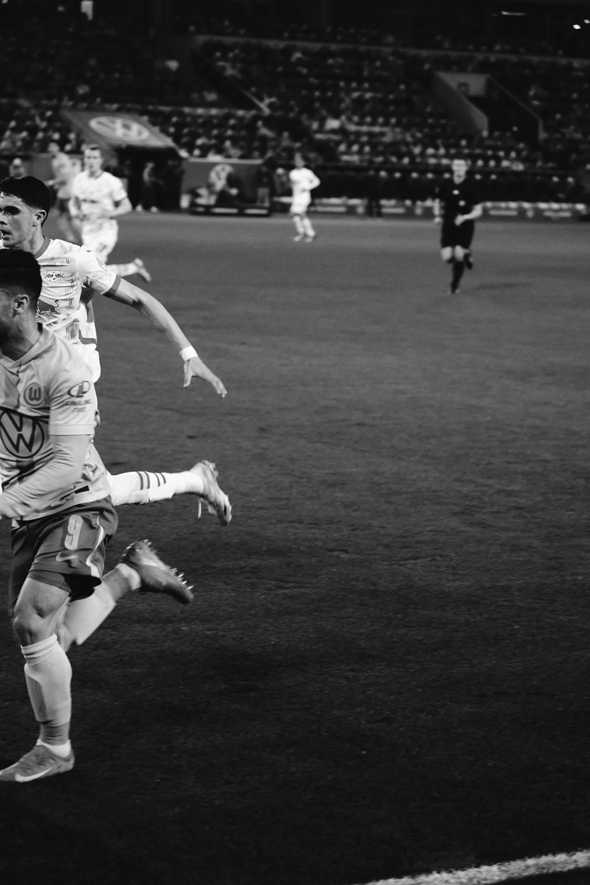 Bundesliga, players sprinting at the edge of the frame, referee in the background, black and white under floodlights, sports photography, photographed by Kilian Amrehn