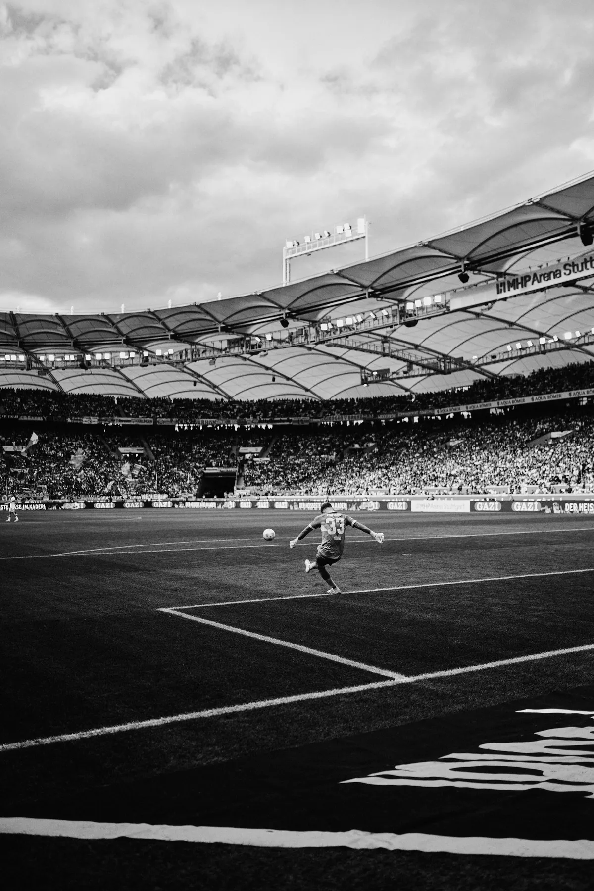 Bundesliga, player taking a corner beneath a curved stadium roof and cloudy sky, black and white, sports photography, photographed by Kilian Amrehn