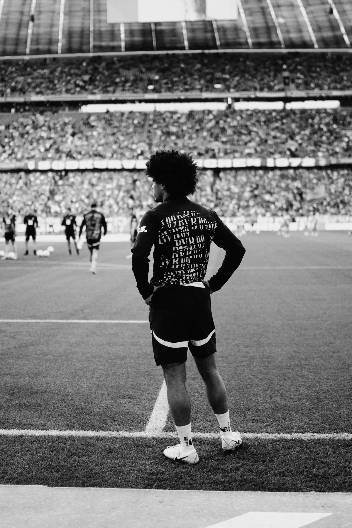 Bundesliga, curly-haired player standing hands on hips at the touchline, back view before full stands, black and white, sports photography, photographed by Kilian Amrehn