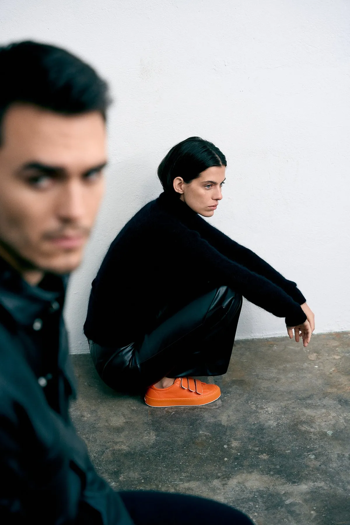 Copenhagen Studios, model in black crouching in orange shoes by a white wall, blurred male face in the foreground, campaign October 2022, photographed by Kilian Amrehn