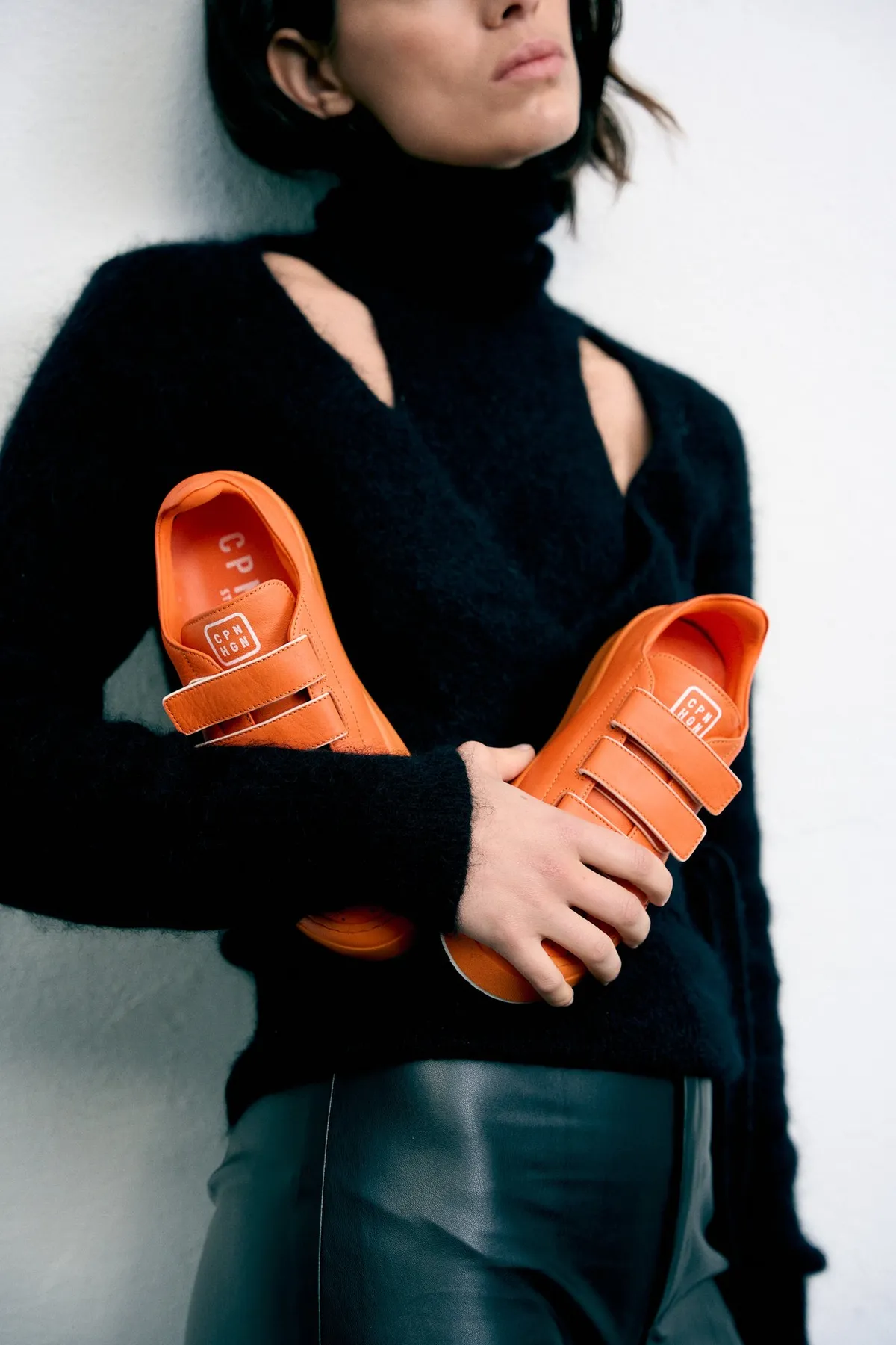 Copenhagen Studios, model in a black cut-out knit holding orange velcro sneakers, leather trousers, campaign October 2022, photographed by Kilian Amrehn