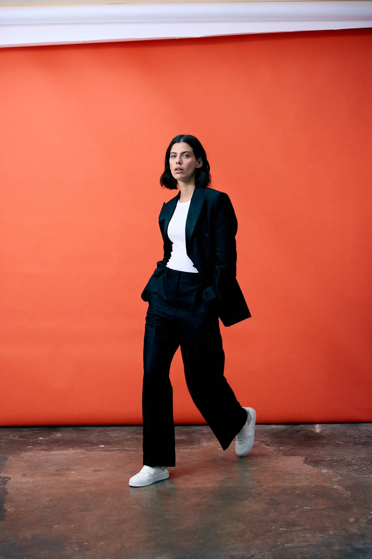 Copenhagen Studios, model in a black suit and white sneakers walking in front of a bright orange backdrop, campaign October 2022, photographed by Kilian Amrehn