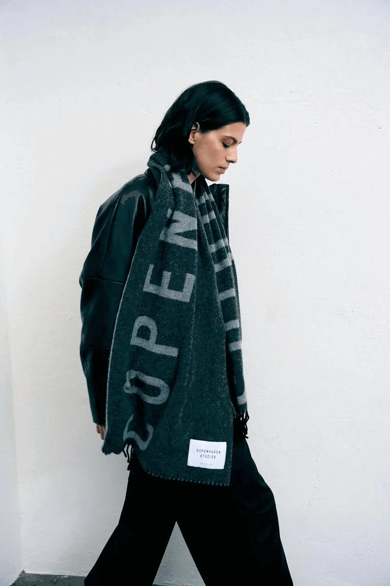 Copenhagen Studios, model in a leather jacket with grey logo scarf striding past a white wall, eyes lowered, campaign October 2022, photographed by Kilian Amrehn