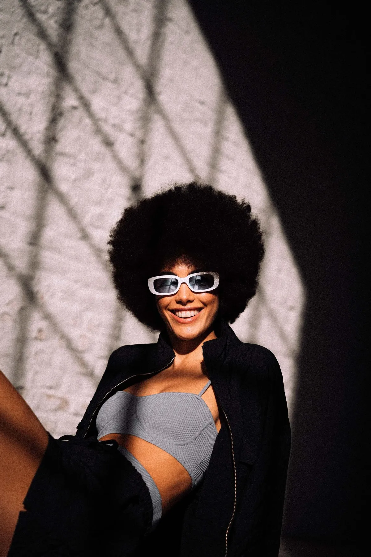 Copenhagen Studios, laughing model with an afro and white sunglasses in a grey bralette, hard lattice shadow on the wall, campaign February 2021, photographed by Kilian Amrehn