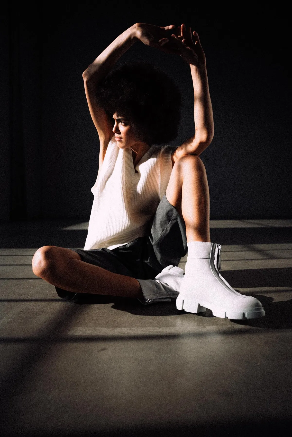 Copenhagen Studios, model with an afro seated on the floor in white sock boots, arms overhead, low light with striped shadows, campaign February 2021, photographed by Kilian Amrehn
