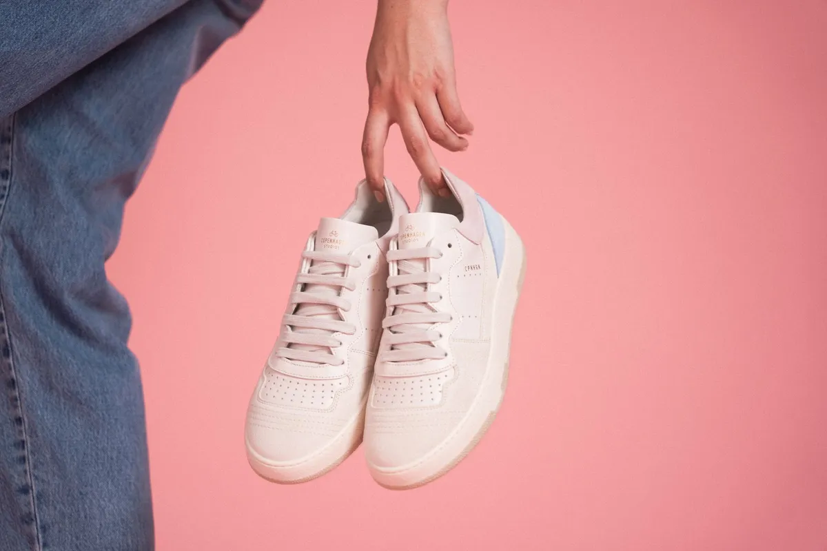 Copenhagen Studios, hand dangling a pair of pale leather sneakers against a pink background, denim leg in frame, campaign June 2021, photographed by Kilian Amrehn