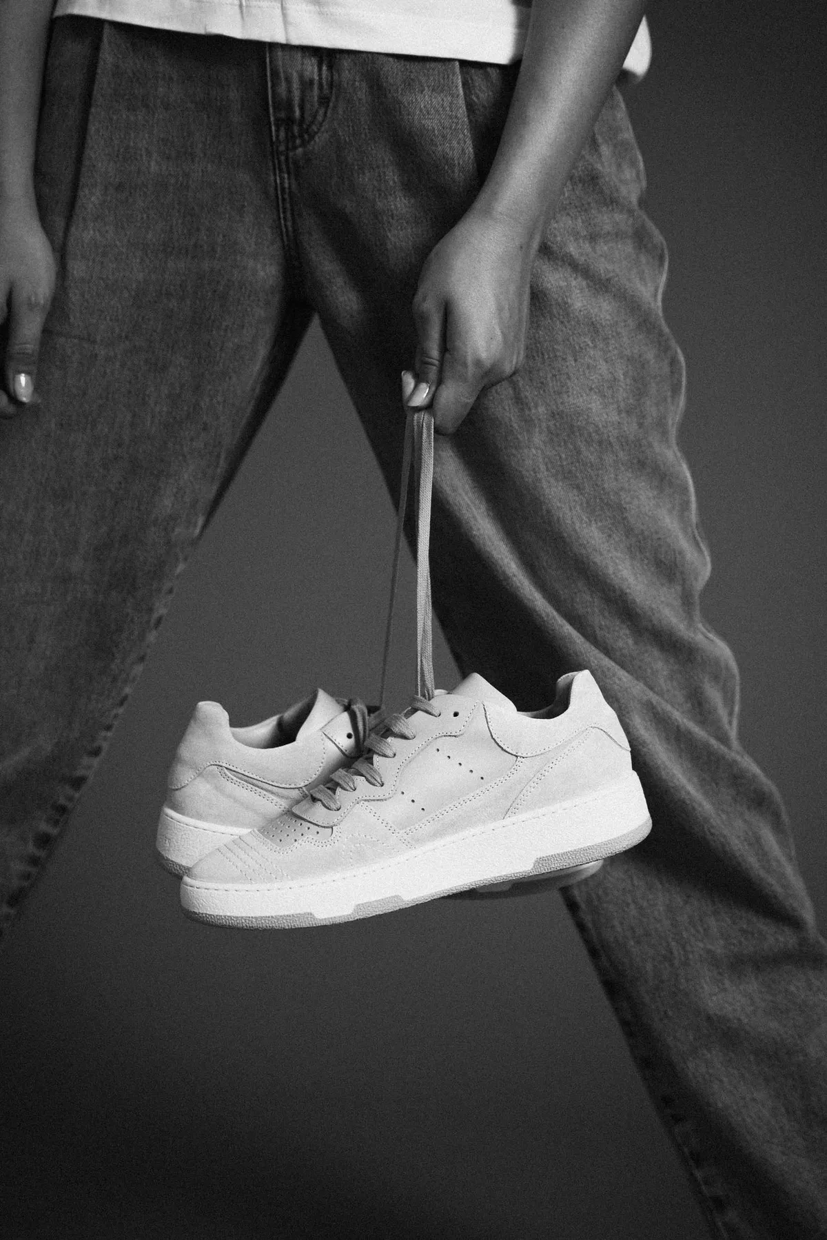 Copenhagen Studios, wide jeans and a pair of white sneakers held by the laces, black and white against a dark background, campaign June 2021, photographed by Kilian Amrehn