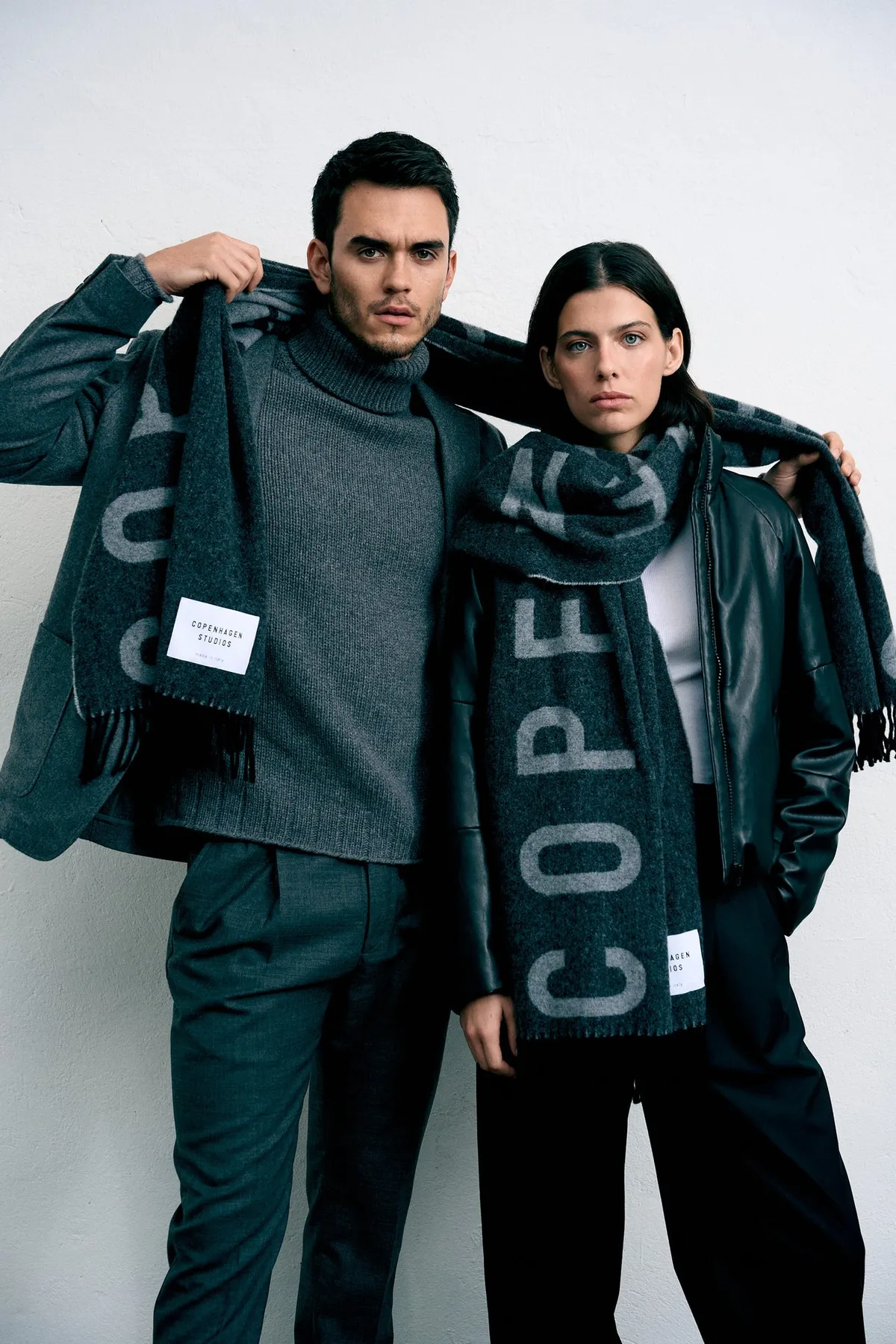 Copenhagen Studios, two models in dark winter clothing sharing a grey logo scarf, light studio background, campaign October 2022, photographed by Kilian Amrehn